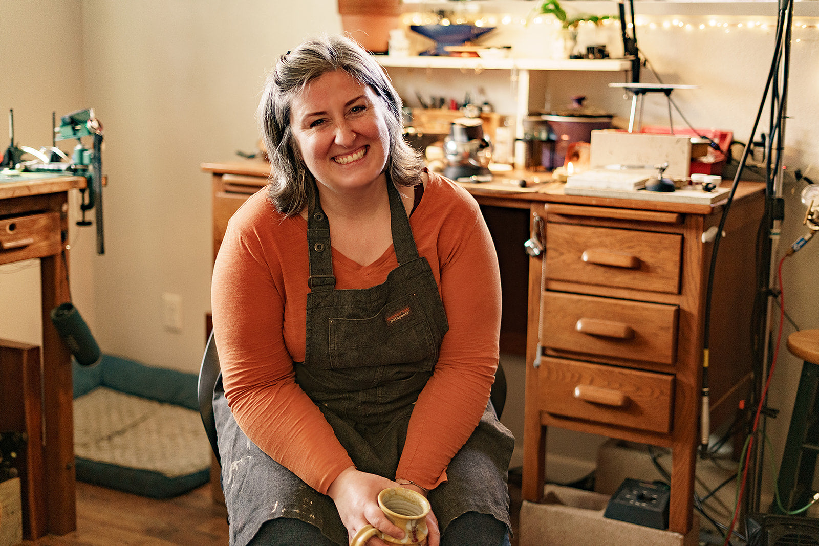 Woman in an apron sitting in a workshop holding a mug